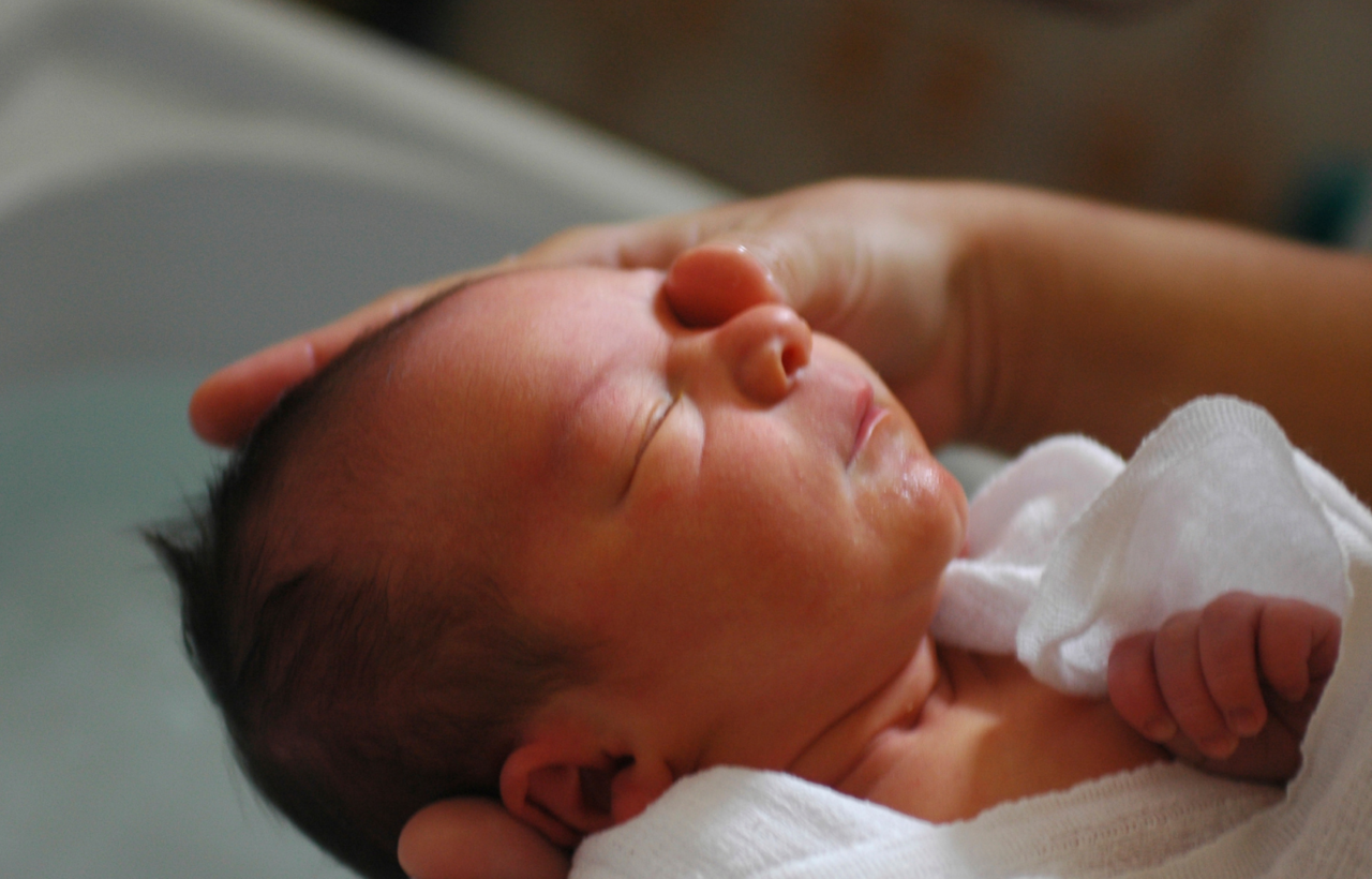 Newborn baby wrapped in a white cloth with an adult hand resting gently on their forehead.