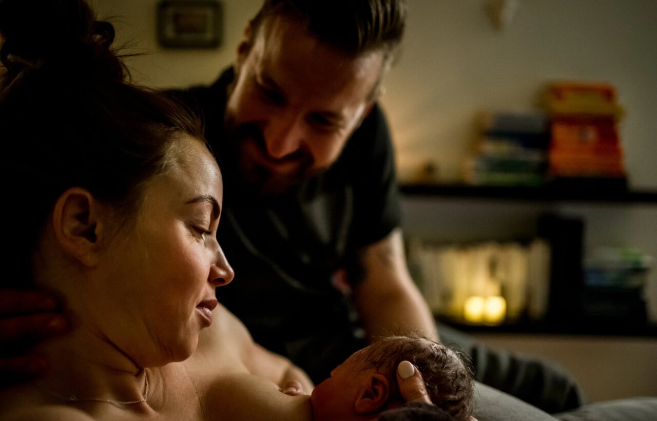 Parent breastfeeds a newborn while another adult leans close in a dimly lit room.