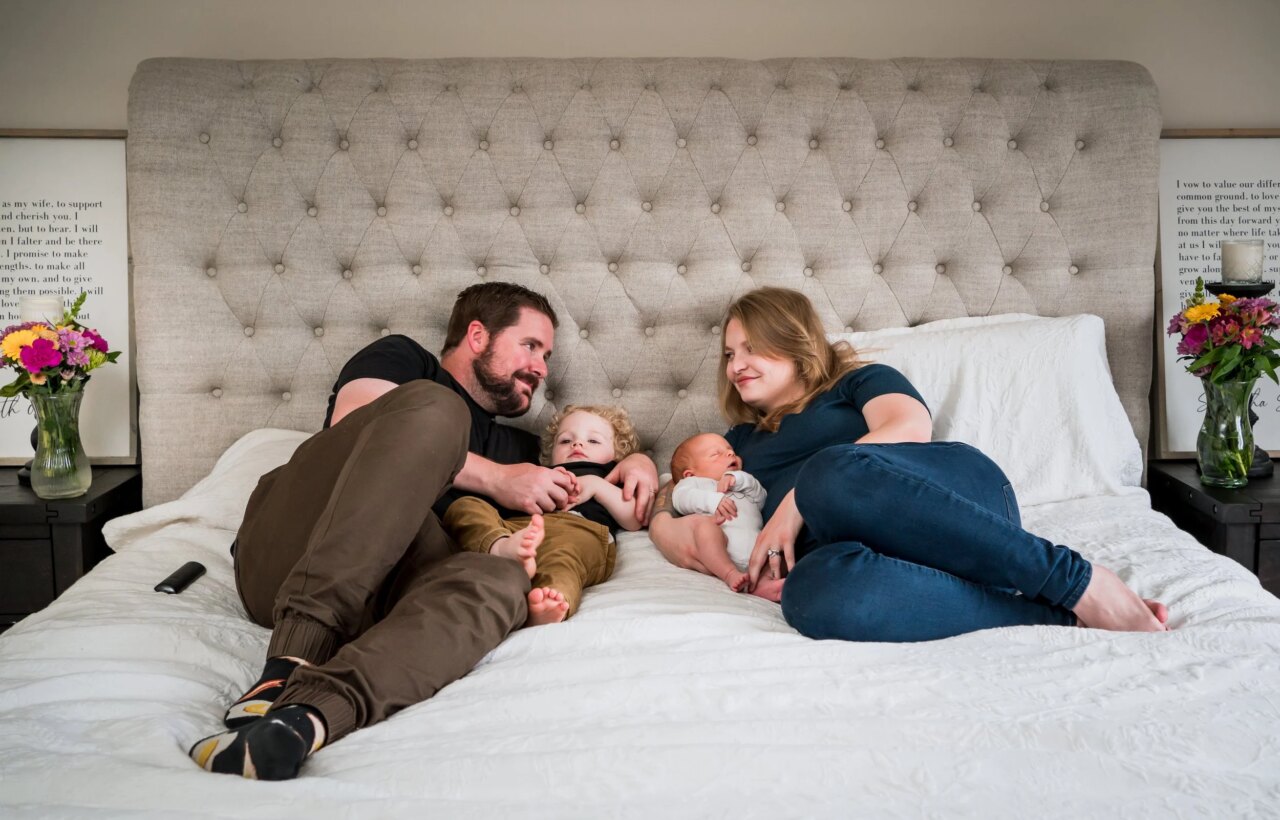 Two adults and two young children lying on a bed in front of a tufted gray headboard.