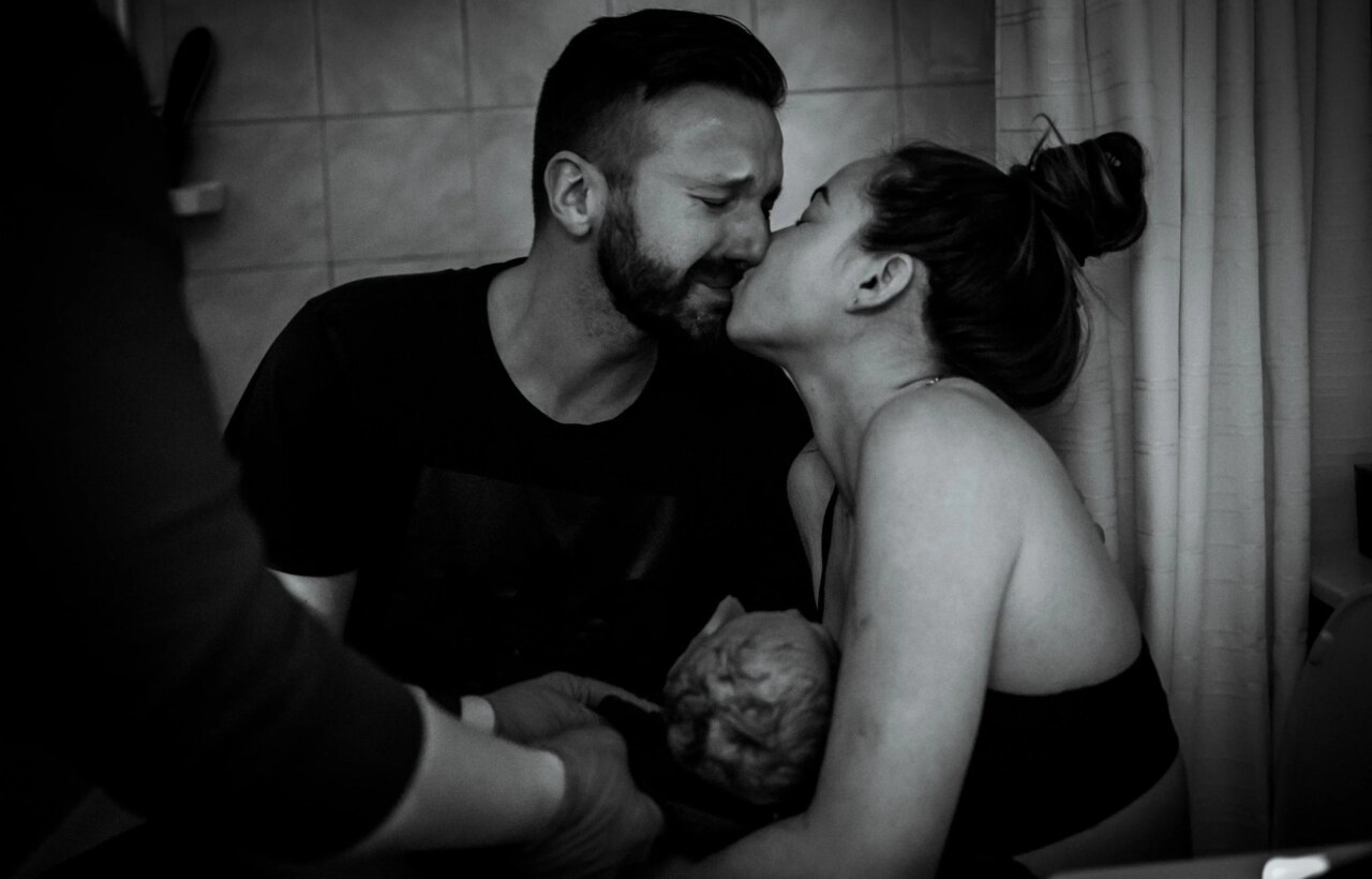Black-and-white scene of two adults close together beside a newborn in a tiled room.
