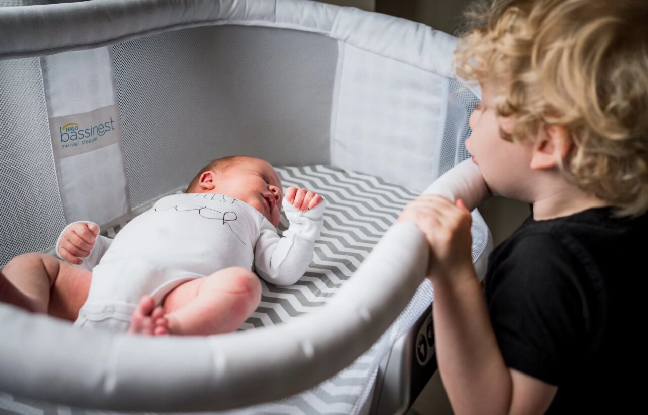 Newborn lying in a bassinet while a young child stands beside it looking in