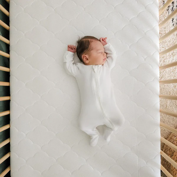Infant in a white sleeper lying on a white quilted crib mattress with wooden crib rails.