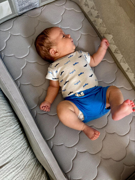 Baby lying on a gray patterned mattress wearing a white shirt and blue shorts.