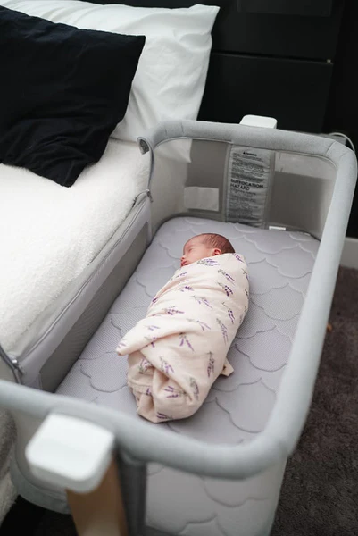 Swaddled baby sleeping in a gray bedside bassinet next to a white bed