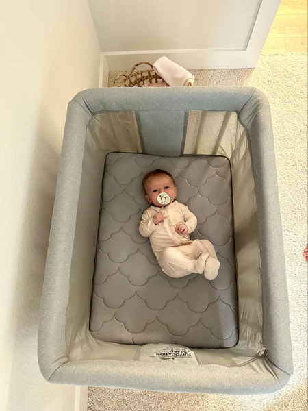 Baby lying in a mesh-sided bassinet on a gray quilted mattress with a pacifier