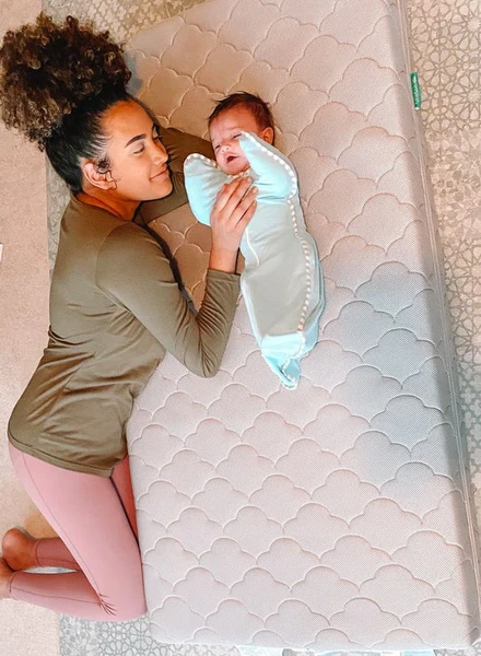 Woman lying beside a crying baby swaddled on a white patterned mattress