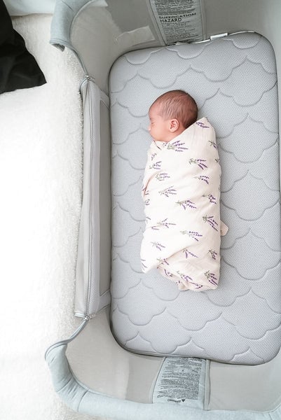 Swaddled baby lying on a gray quilted mattress inside a light-colored bassinet