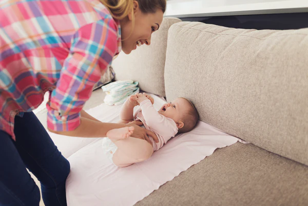Adult in a plaid shirt leans over a baby lying on a white pad on a beige sofa.