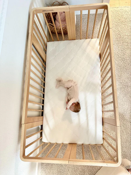 Baby curled on a white quilted mattress inside a light wood crib, viewed from above.