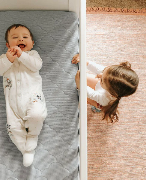 Baby in a white sleeper lying on a crib mattress as a child looks in from beside the crib.