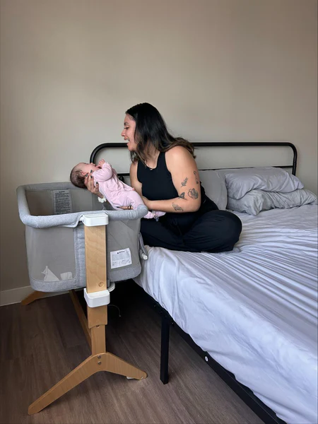 Woman seated on a bed leaning toward a baby in a pink outfit lying in a bedside bassinet.
