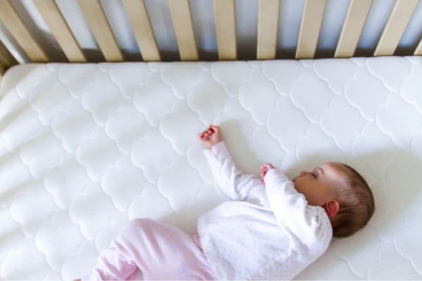Baby lying on a white quilted crib mattress beside wooden crib slats