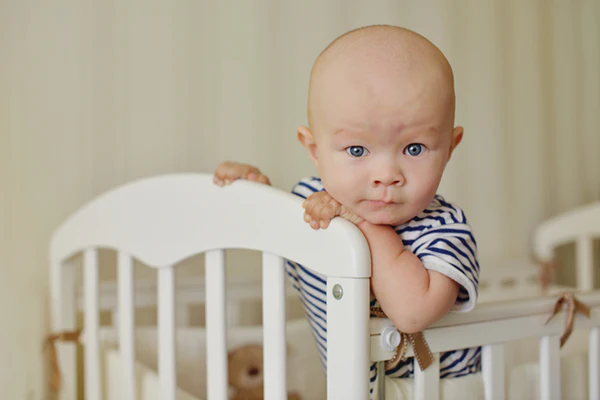 Baby in a striped shirt leaning over the rail of a white crib