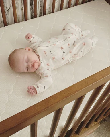 Baby lying on their back in a wooden crib on a light-colored mattress.