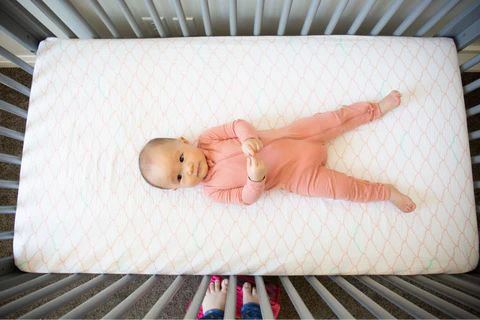 Baby in a pink sleeper lying on a crib mattress, with crib rails and adult feet visible at the bottom.