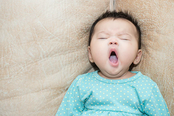 Baby in a teal polka dot outfit yawning with eyes closed on beige fabric
