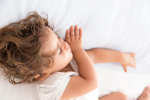 Young child with curly hair lying on white bedding, viewed from above.