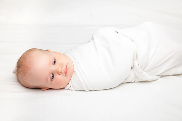 Swaddled baby lying on a white bed and looking toward the camera.