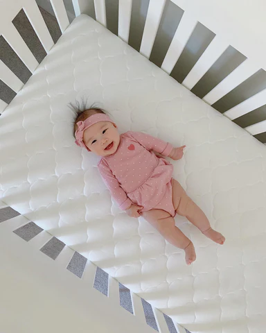 Smiling baby in a pink outfit lying on a white crib mattress
