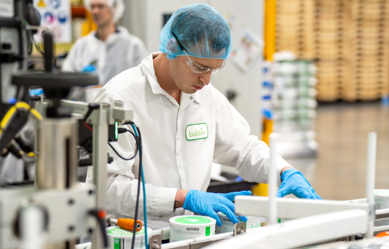 Worker in hairnet, safety glasses, and blue gloves handles bobbie containers on a production line.