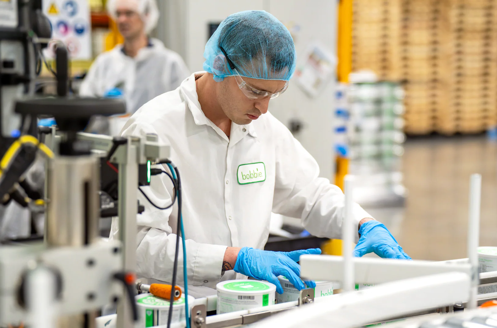 Worker in hairnet, safety glasses, and blue gloves handles bobbie containers on a production line.
