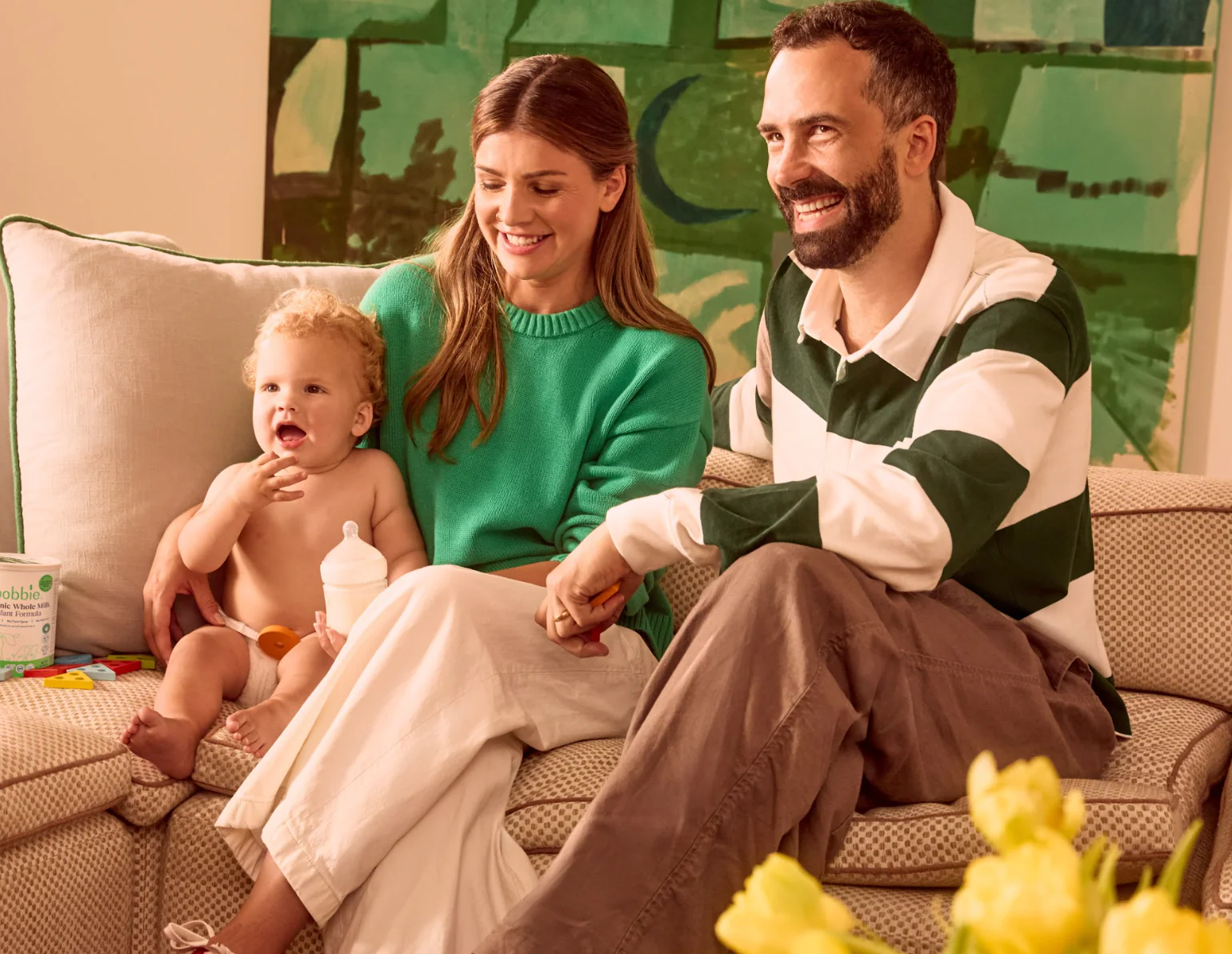 Two adults sit on a couch with a smiling baby holding a bottle, beside a partly visible Bobbie formula can.