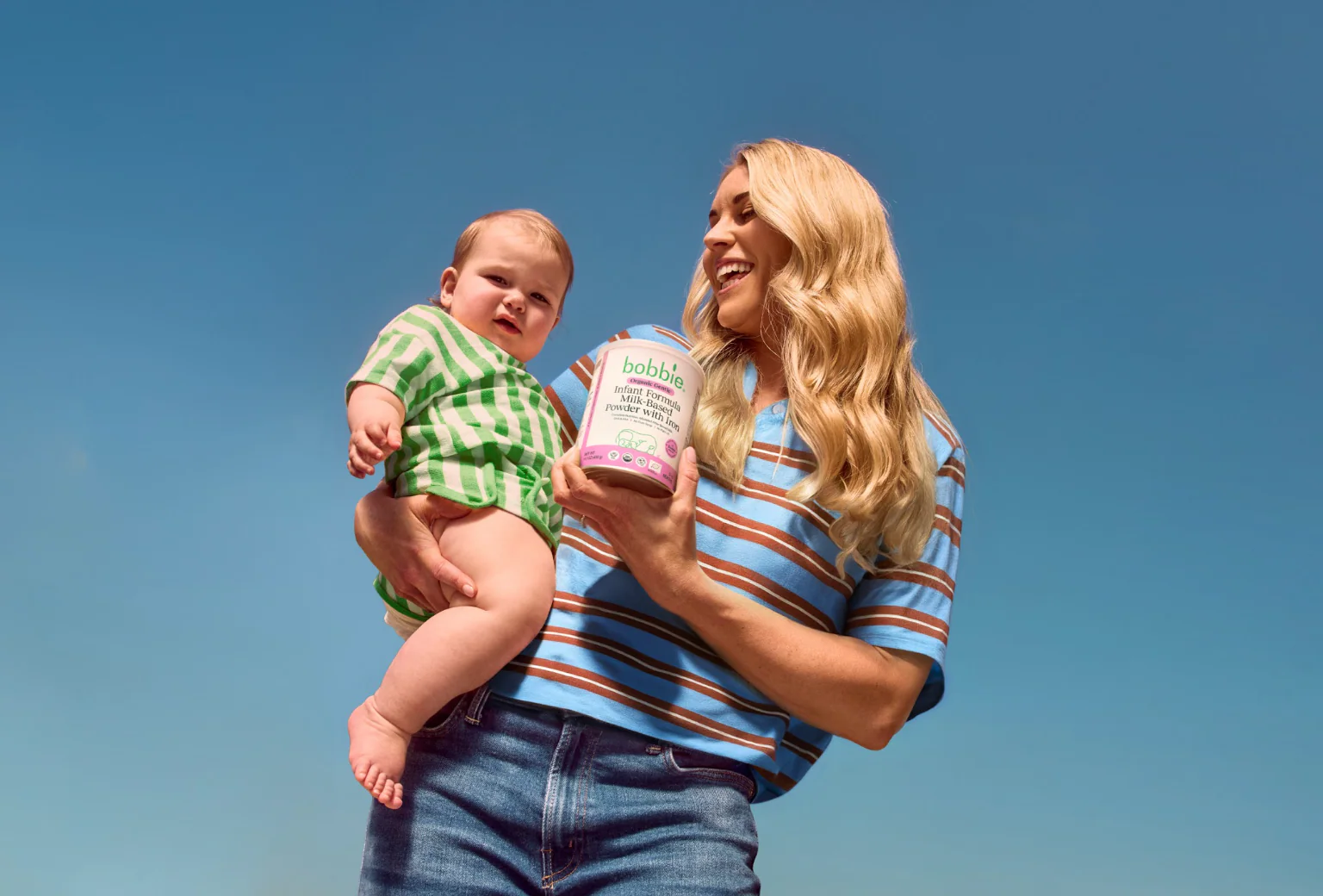 Smiling adult holding a baby and a can of Bobbie organic infant formula against a blue background.