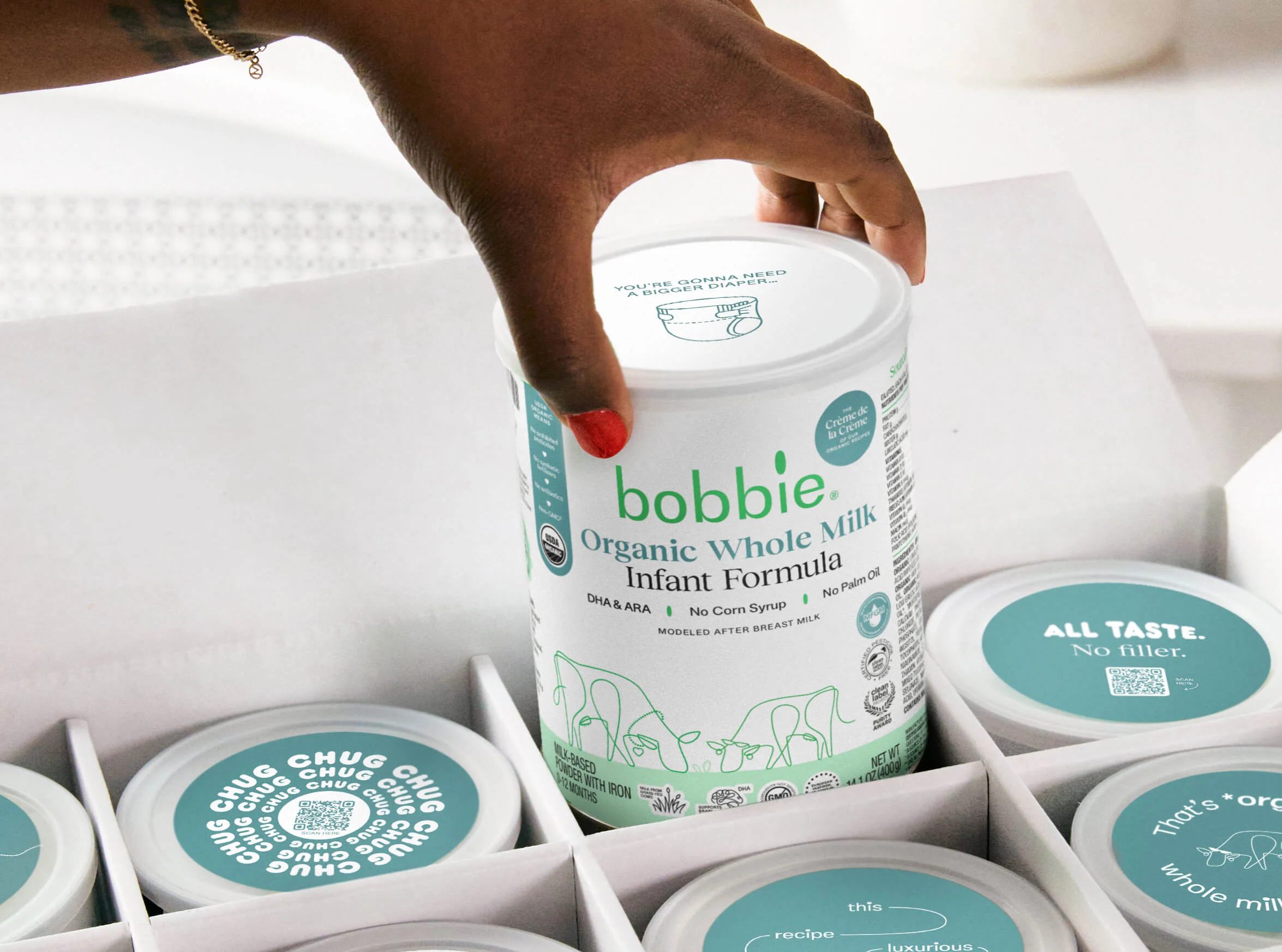 Hand lifting a Bobbie Organic Whole Milk Infant Formula can from a box with other teal-lidded cans