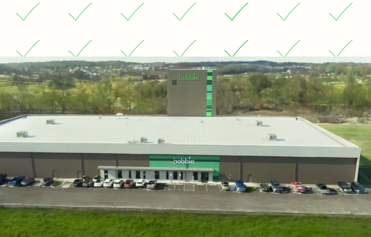 Aerial view of a large building with bobbie. signage, parked cars, trees, fields, and green check marks above.