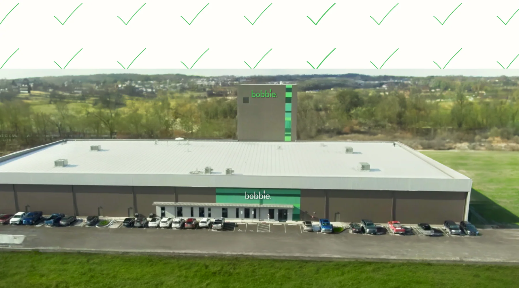 Aerial view of a large building with bobbie. signage, parked cars, trees, fields, and green check marks above.