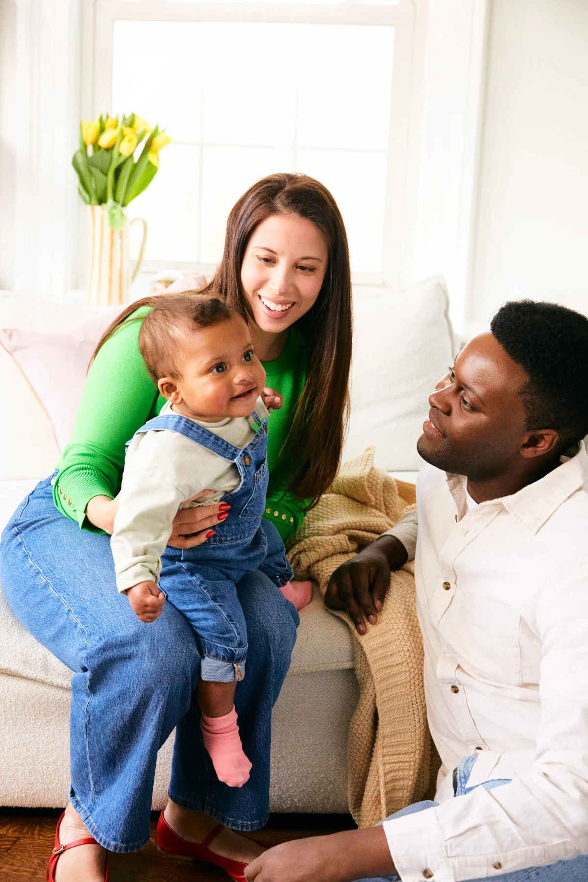 Two adults smiling with a baby on a couch in a bright room with yellow flowers by the window.