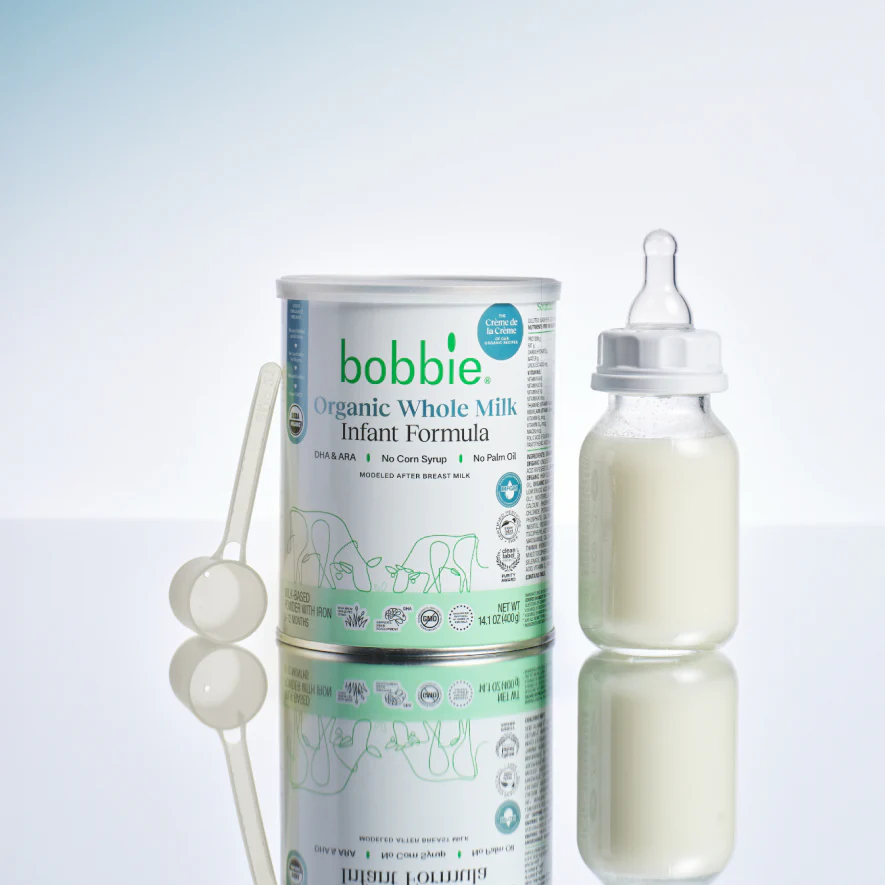 Bobbie Organic Whole Milk Infant Formula can beside a filled baby bottle and measuring scoops.