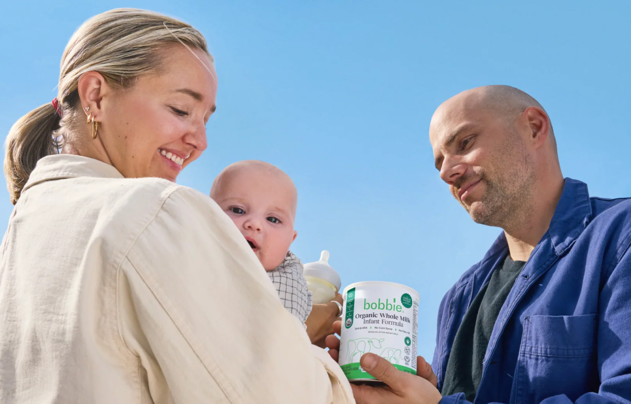 Two adults with a baby holding a Bobbie infant formula can outdoors under a blue sky.