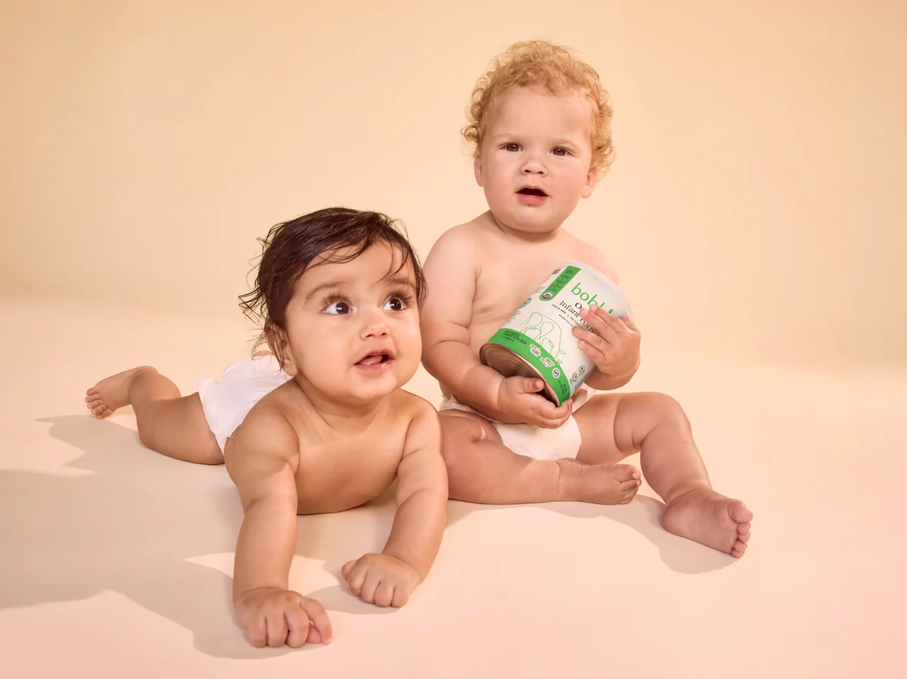 Two babies in diapers on a peach backdrop, one holding a Bobbie infant formula can.