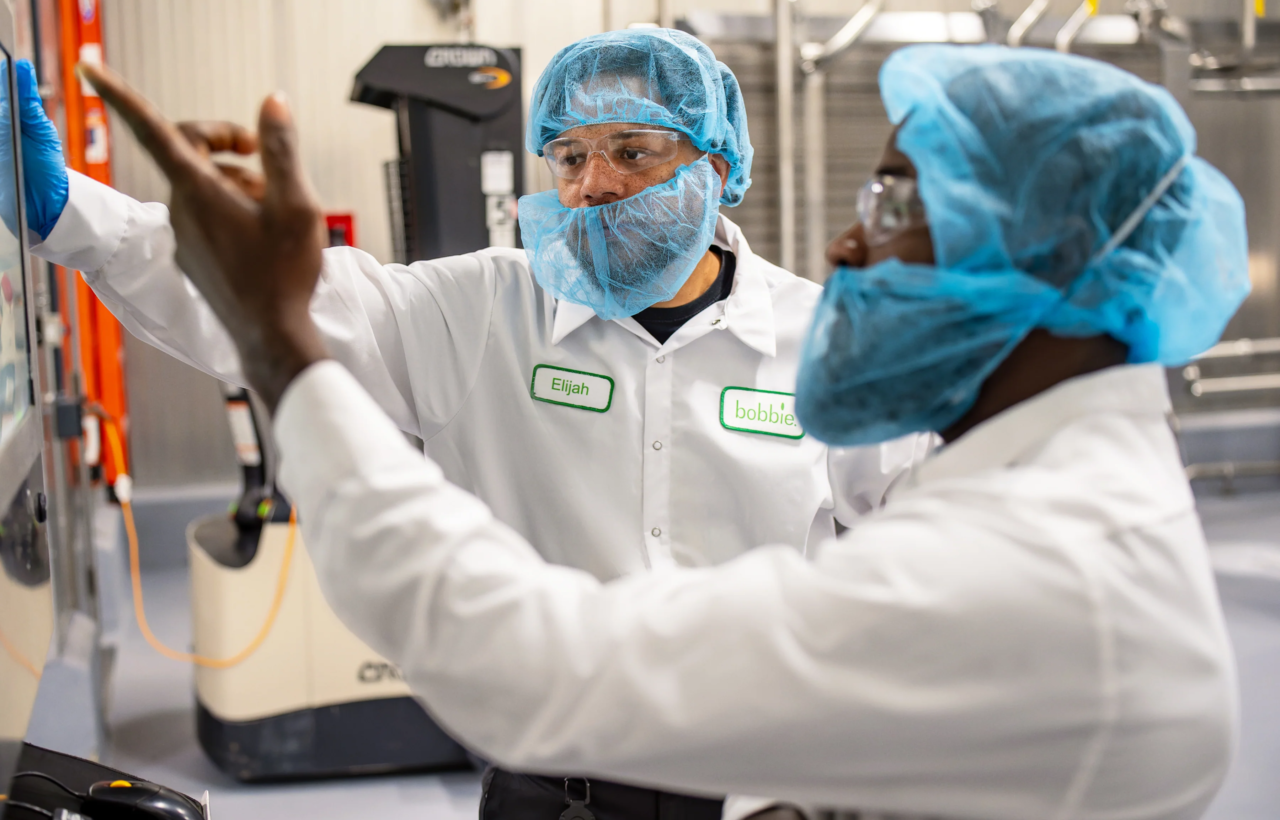 Two workers in hair nets and lab coats review a screen in a production facility.
