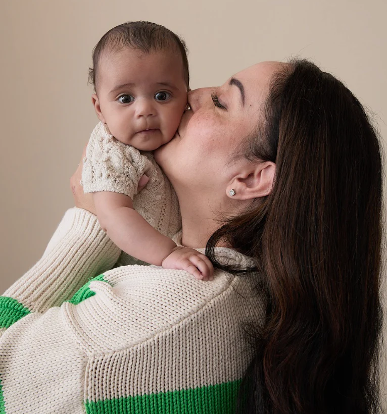 Woman holding a baby and kissing the baby's cheek against a plain beige background.
