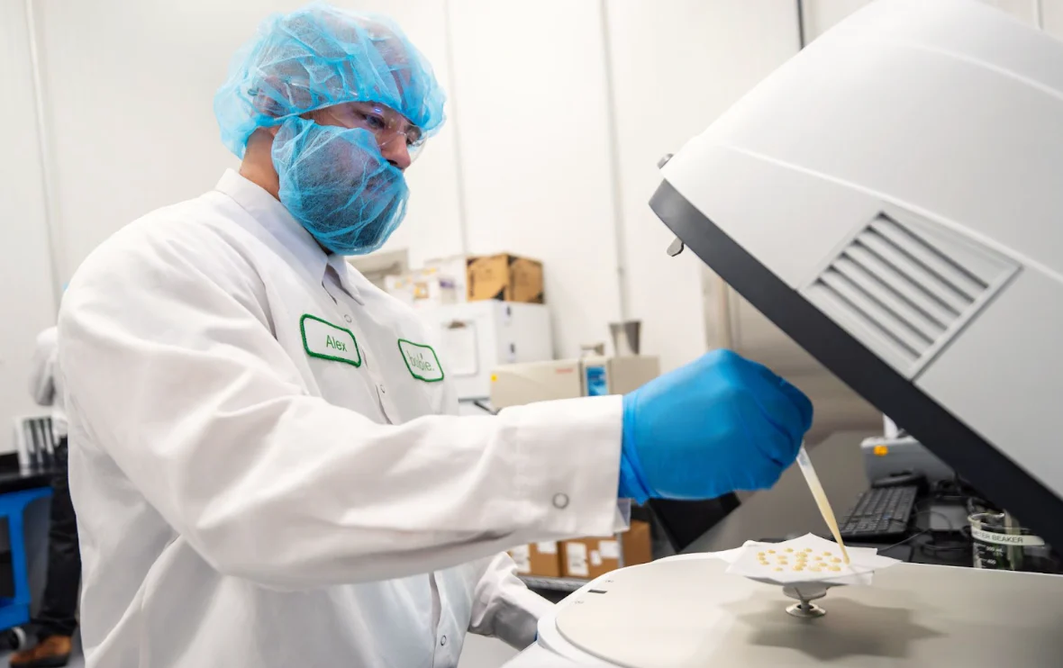 Worker in protective lab gear handles small samples on white paper beside equipment.