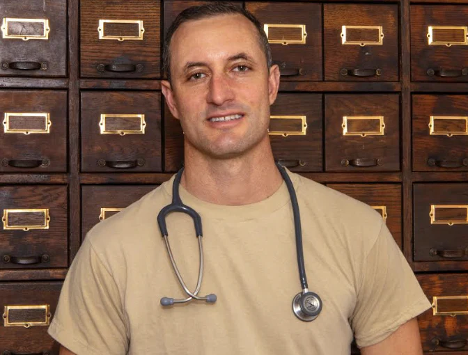 Man wearing a stethoscope stands in front of wooden drawers with brass label holders.