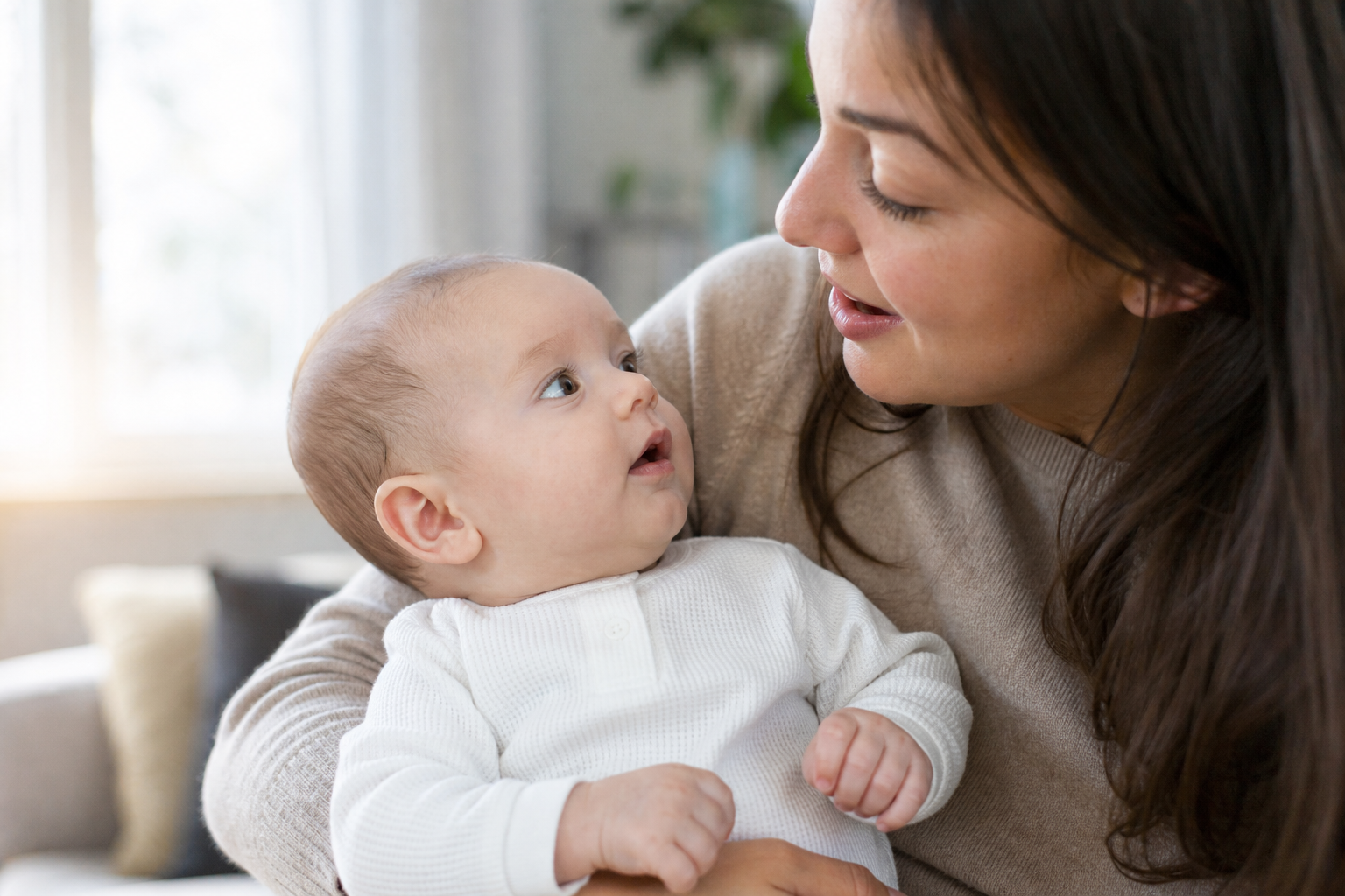 An alert baby turns toward a caregiver speaking nearby in a softly lit home.