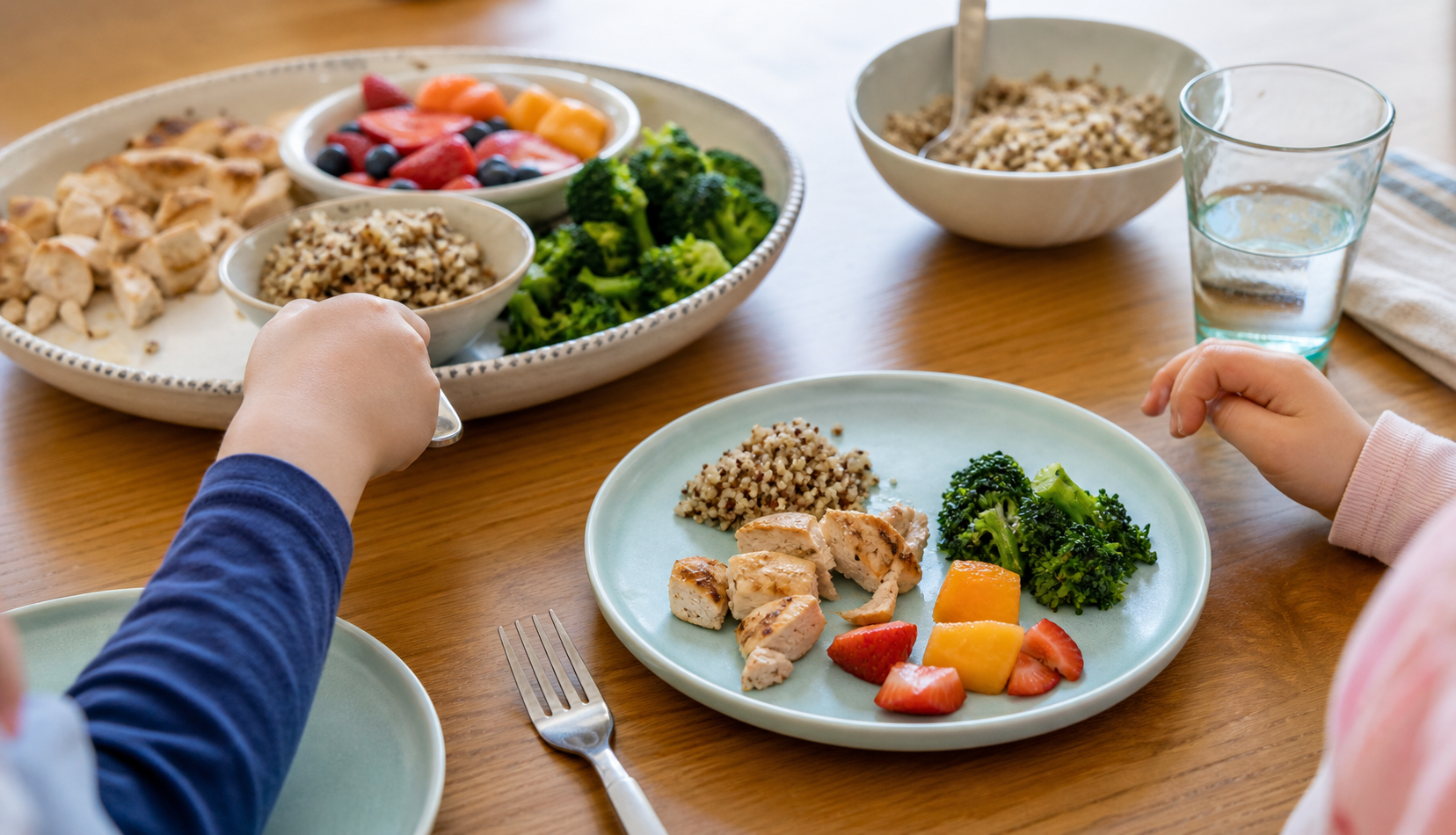 A small child-sized plate with modest portions of grains, protein, vegetables, and fruit beside extra food on a serving platter.