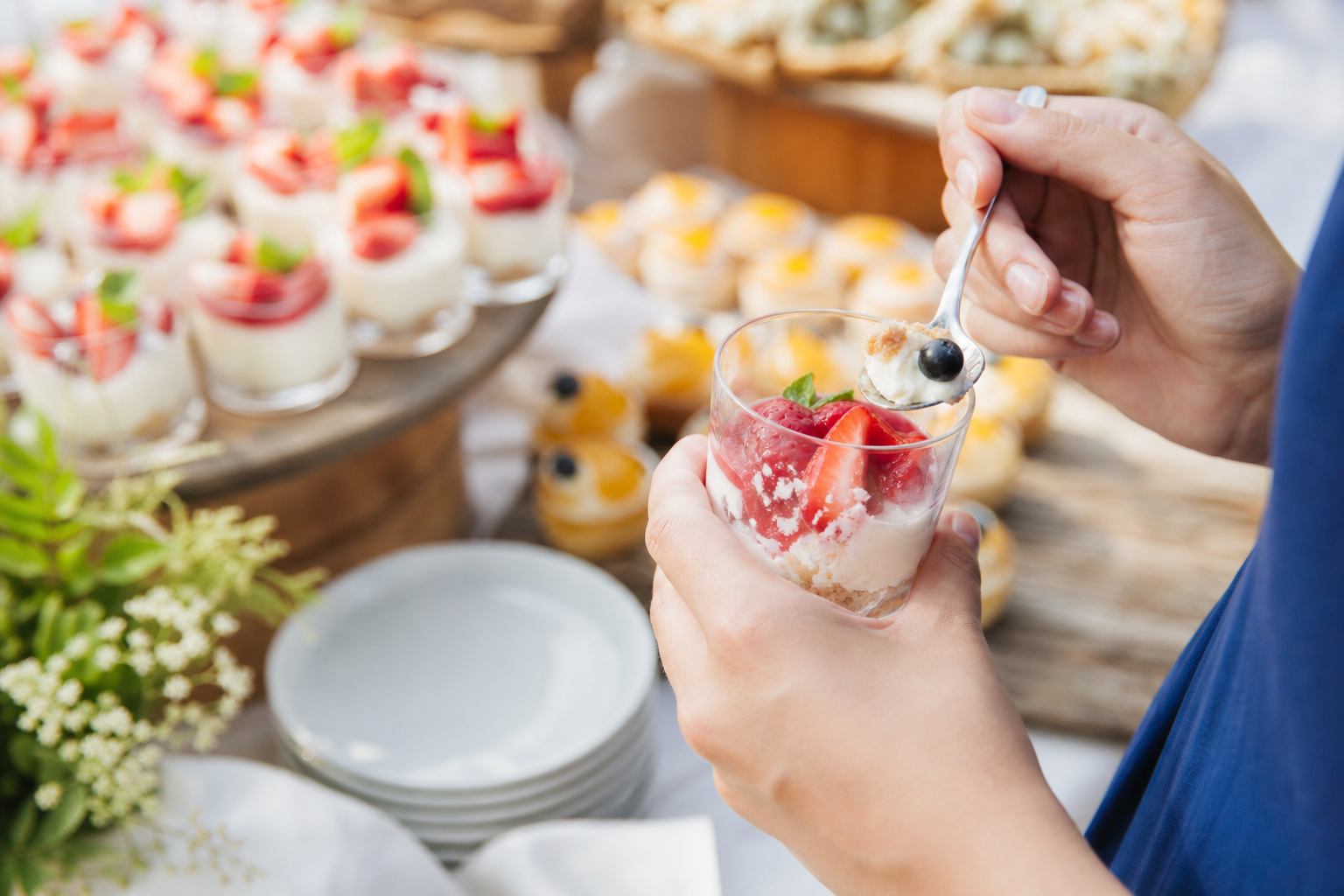 A guest holds a small dessert cup and spoon beside an outdoor buffet with bite-size desserts in the background.