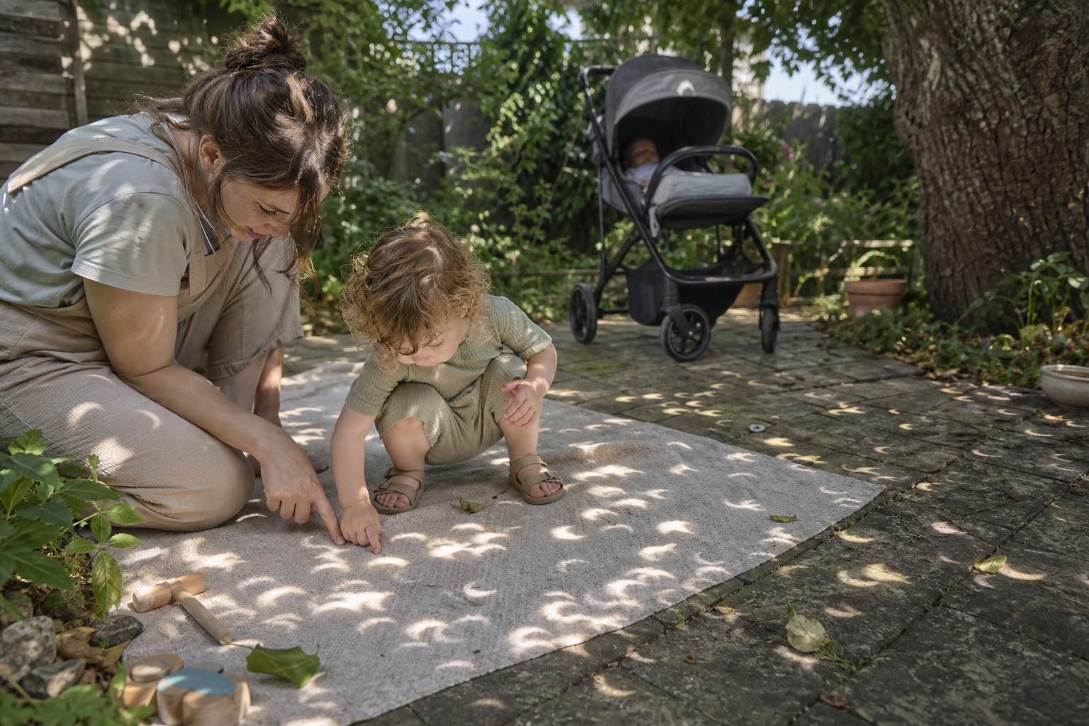 A supervised toddler plays with crescent-shaped patches of projected sunlight beneath a leafy tree while a baby rests in a shaded pram.