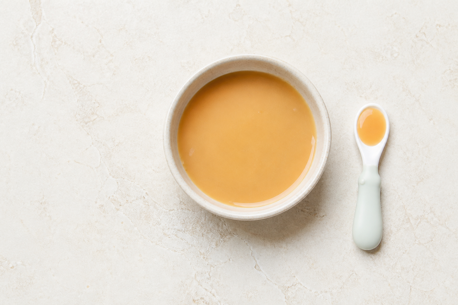 A small bowl holds smooth peanut butter thinned to a loose, spoonable consistency beside a baby spoon.