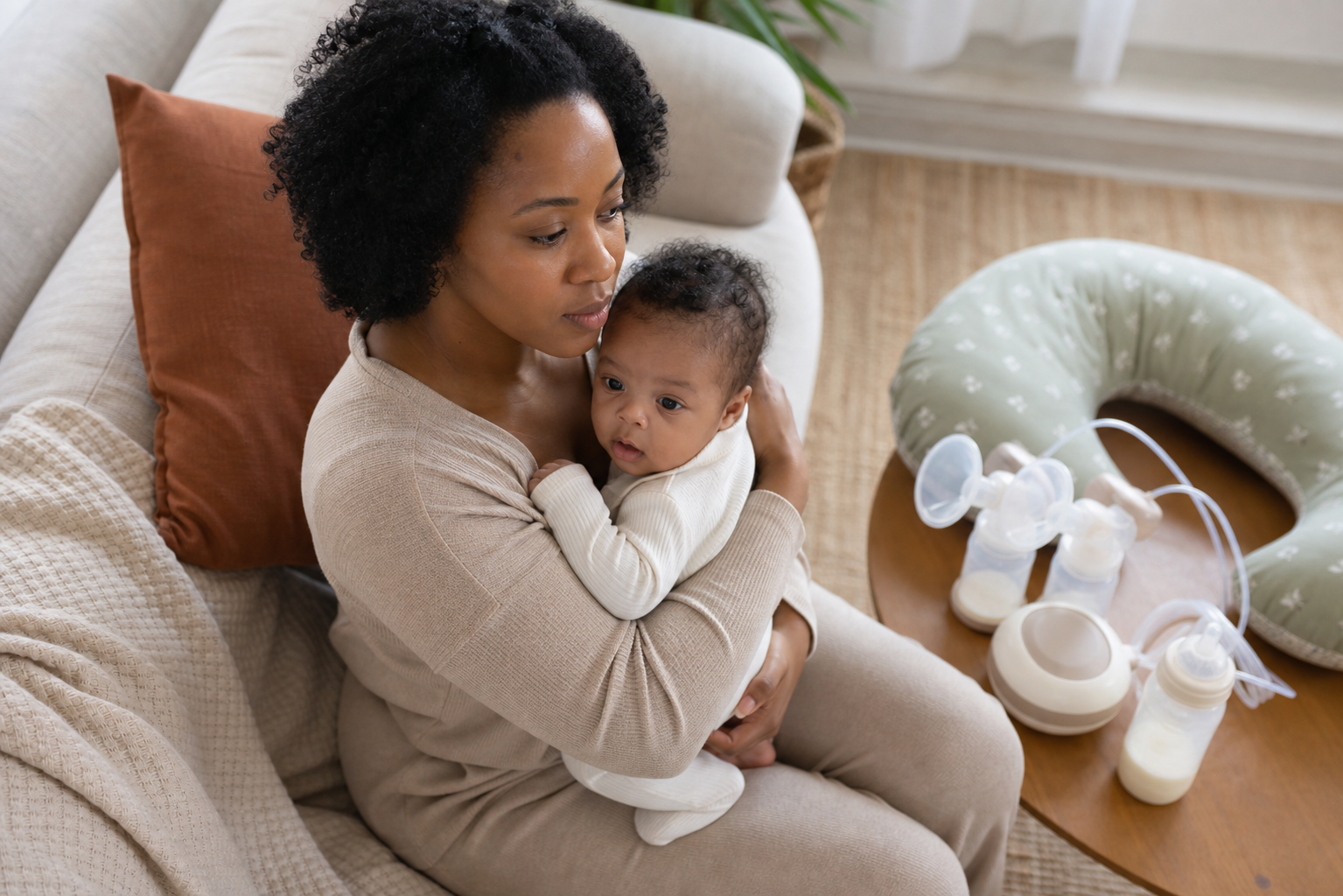 A Black parent holds their baby beside a nursing pillow, breast pump parts, and a prepared bottle.