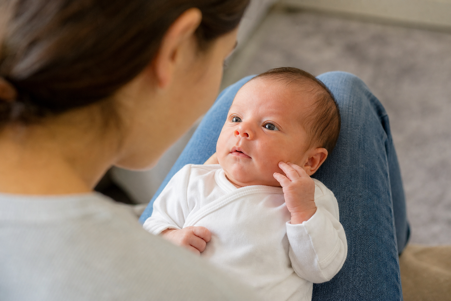 An awake newborn looks toward a nearby caregiver who is speaking softly while supporting the baby on their lap.