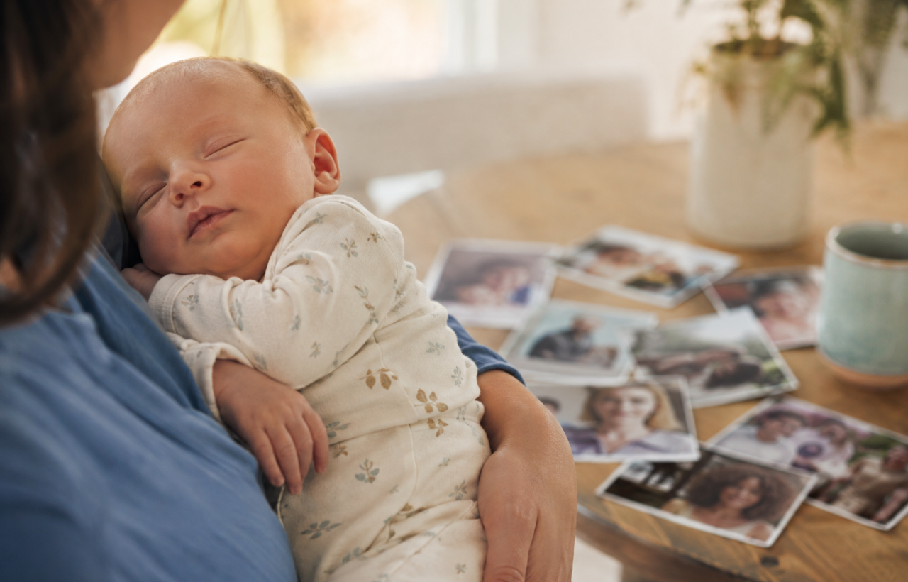 A newborn rests in a caregiver's arms beside softly blurred family photographs showing relatives with varied appearances.