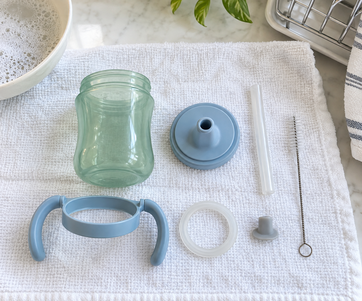 A toddler cup is fully disassembled on a towel, with its lid, straw, valve, gasket, handle ring, and cleaning brush visible.
