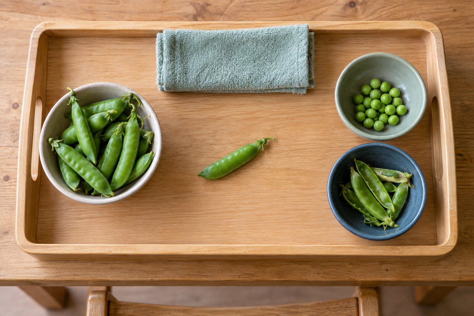 An overhead view of a tray arranged with fresh pea pods, a clear work area, two sorting bowls, and a cleanup cloth.