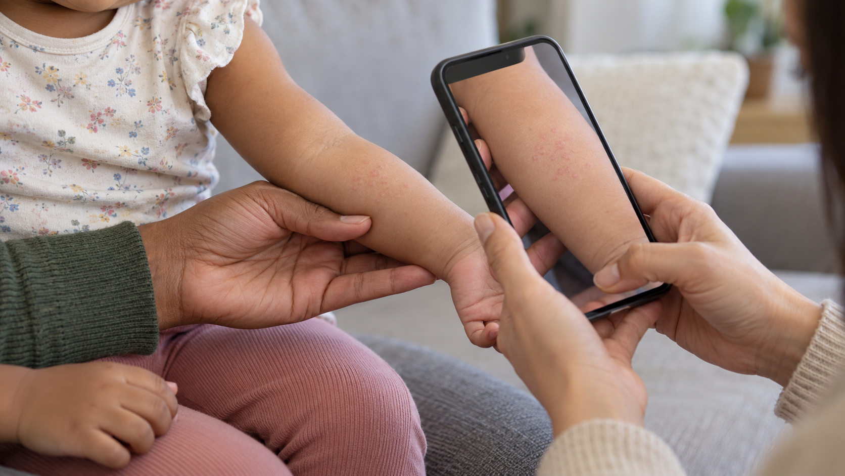 A caregiver photographs a small cluster of pink spots on a toddler's forearm in natural light while another adult gently steadies the arm.